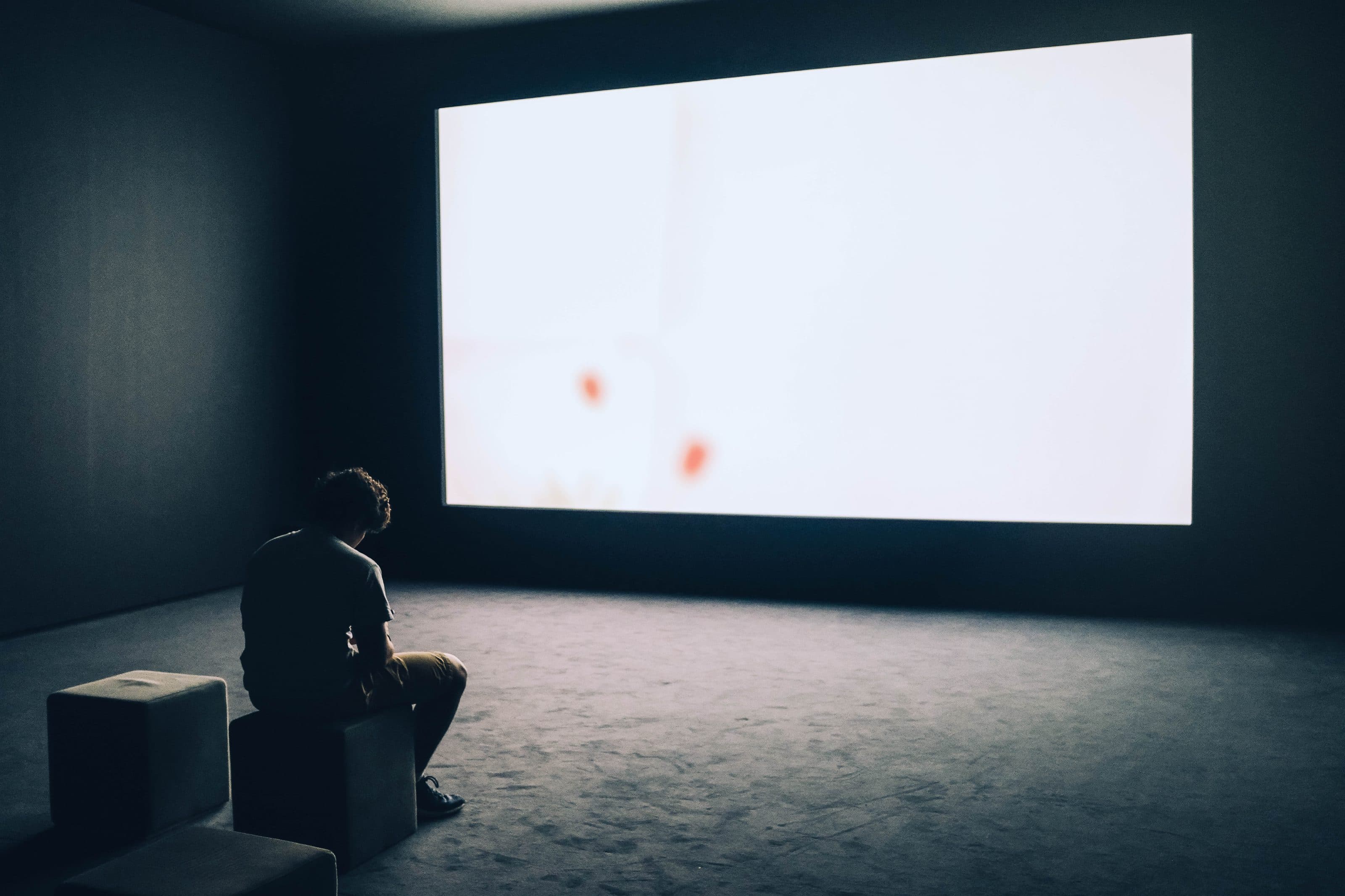 A lone viewer seated in front of a lit projection screen in a darkened room.