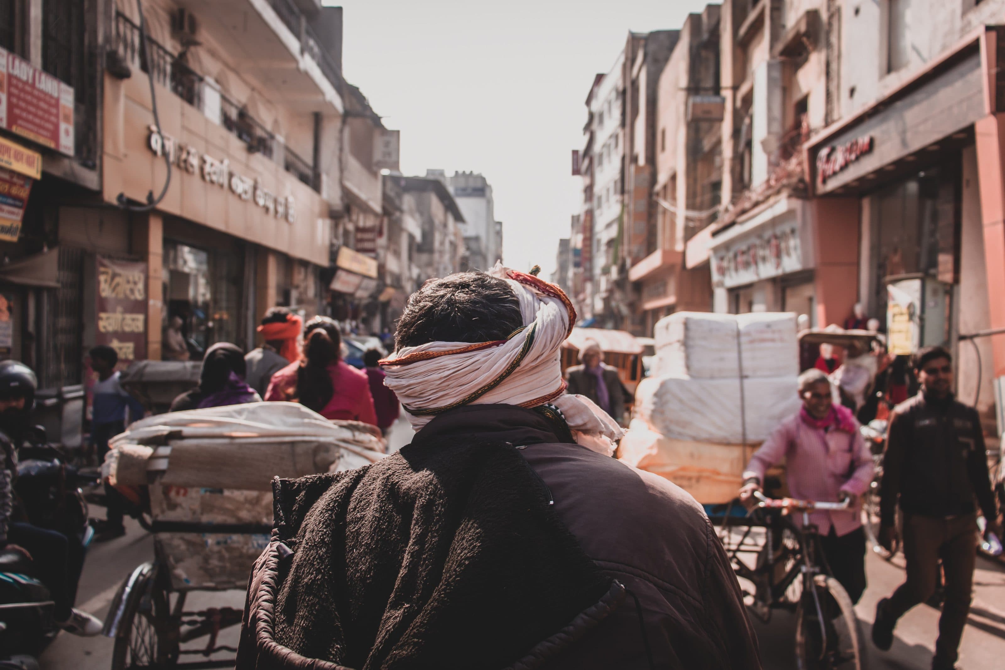 A man in a turban carries goods through a crowded market street in India.