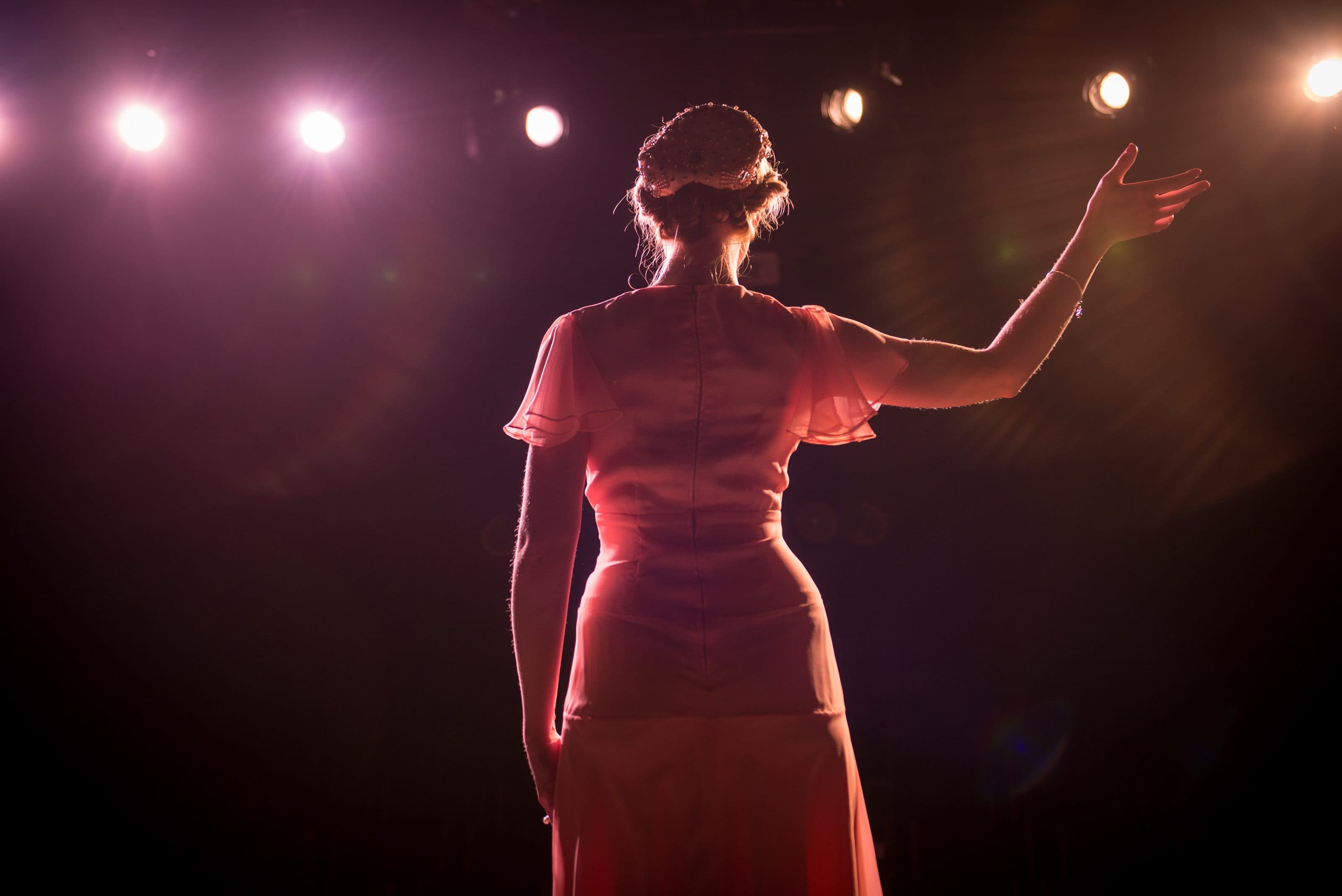 A performer on stage with one arm raised, backlit by theatre lights.
